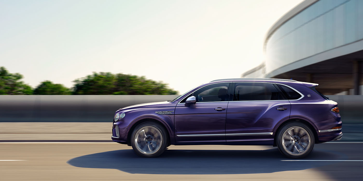 Bentley Riyadh Bentley Bentayga Extended Wheelbase Mulliner luxury SUV profile in Tanzanite Purple paint, driving on an country road past trees.