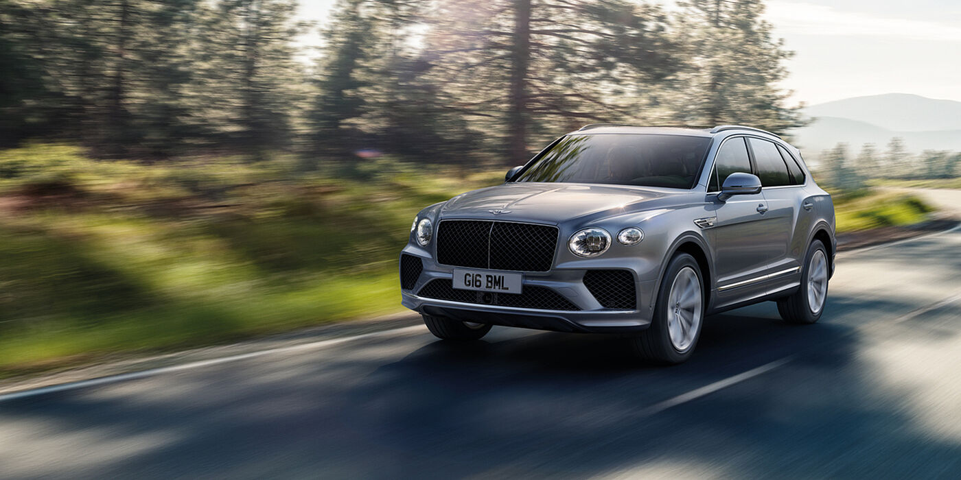 Bentley Riyadh Bentley Bentayga luxury SUV front three quarter in Moonbeam paint, driving on an country road past trees.