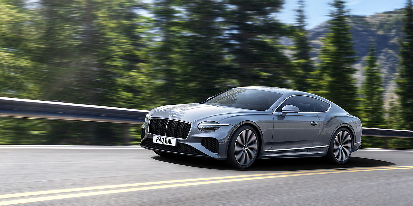 Bentley Riyadh New Bentley Continental GT coupe in Silverlake paint and 22 inch ten-spoke wheels, driving dynamically on a tree-lined road in the sunshine