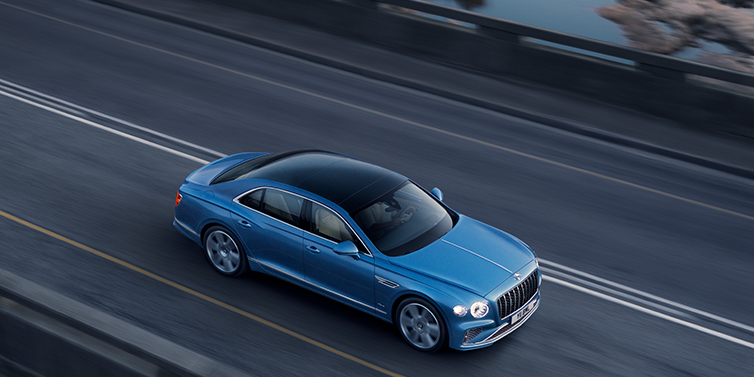 Bentley Riyadh New Bentley Flying Spur Azure sedan in Light windsor blue paint by Mulliner, with 22 inch Azure wheels seen from overhead driving dynamically on a country road