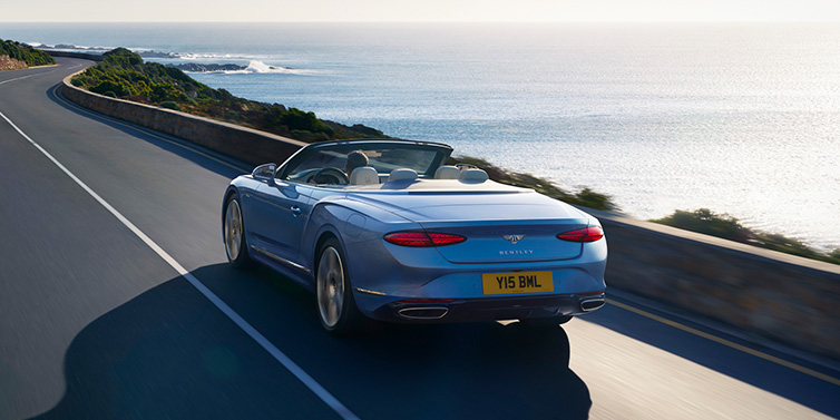Bentley Riyadh New Bentley Continental GTC Azure convertible rear view in Light Windsor blue paint with 22 inch ten spoke wheels driving dynamically on an ocean road in the sunshine