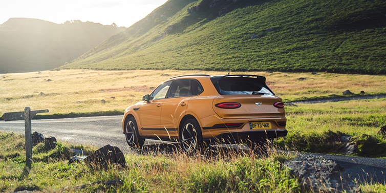 Bentley Riyadh Bentley Bentayga S SUV rear three quarter in Sunburst Gold paint on a road surrounded by green hills