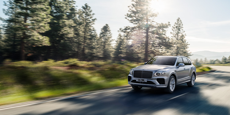 Bentley Riyadh Bentley Bentayga SUV front three quarter in Moonbeam silver paint driving dynamically along a tree-lined road