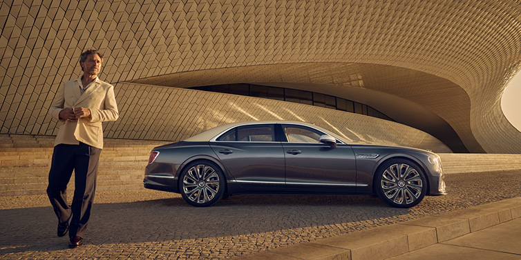 Bentley Riyadh Bentley Flying Spur Mulliner sedan in Rose Gold over Magnetic duotone paint parked by a curved wall with person in suit jacket standing on the left